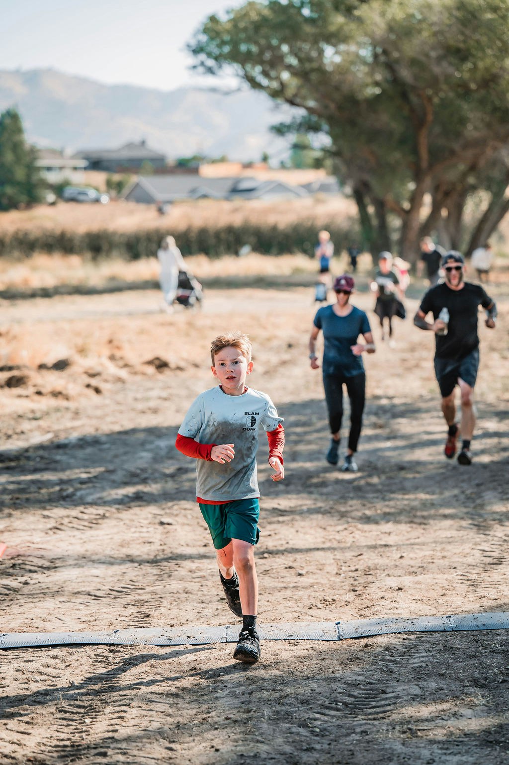 A child runs on a dirt path during a race, with several people running behind, in a sunny outdoor setting.