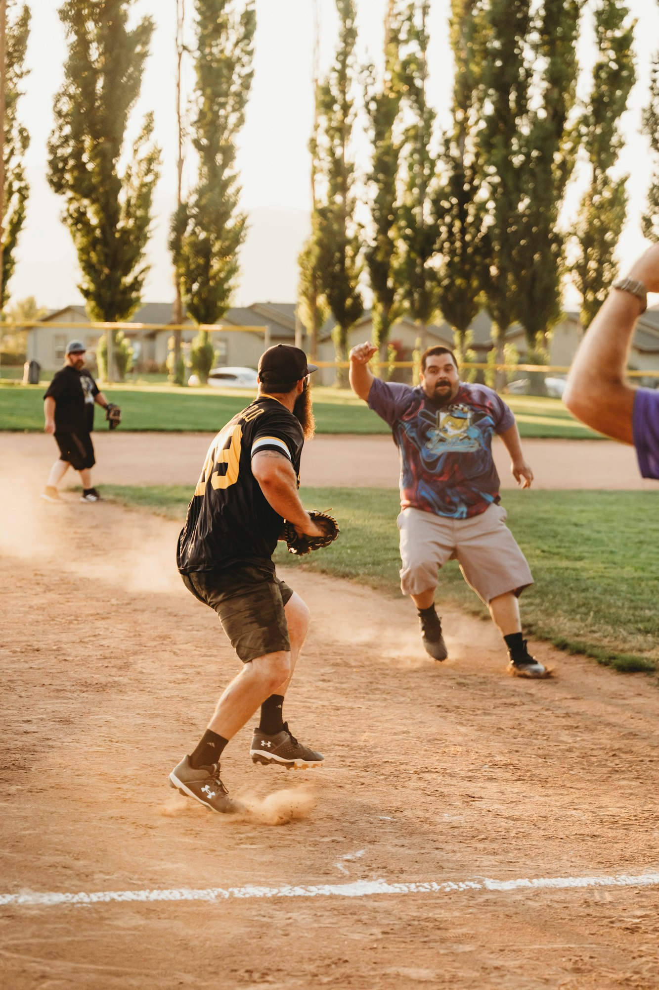 Men playing softball, one slides into base while another attempts a tag. Dust flies, action shot.