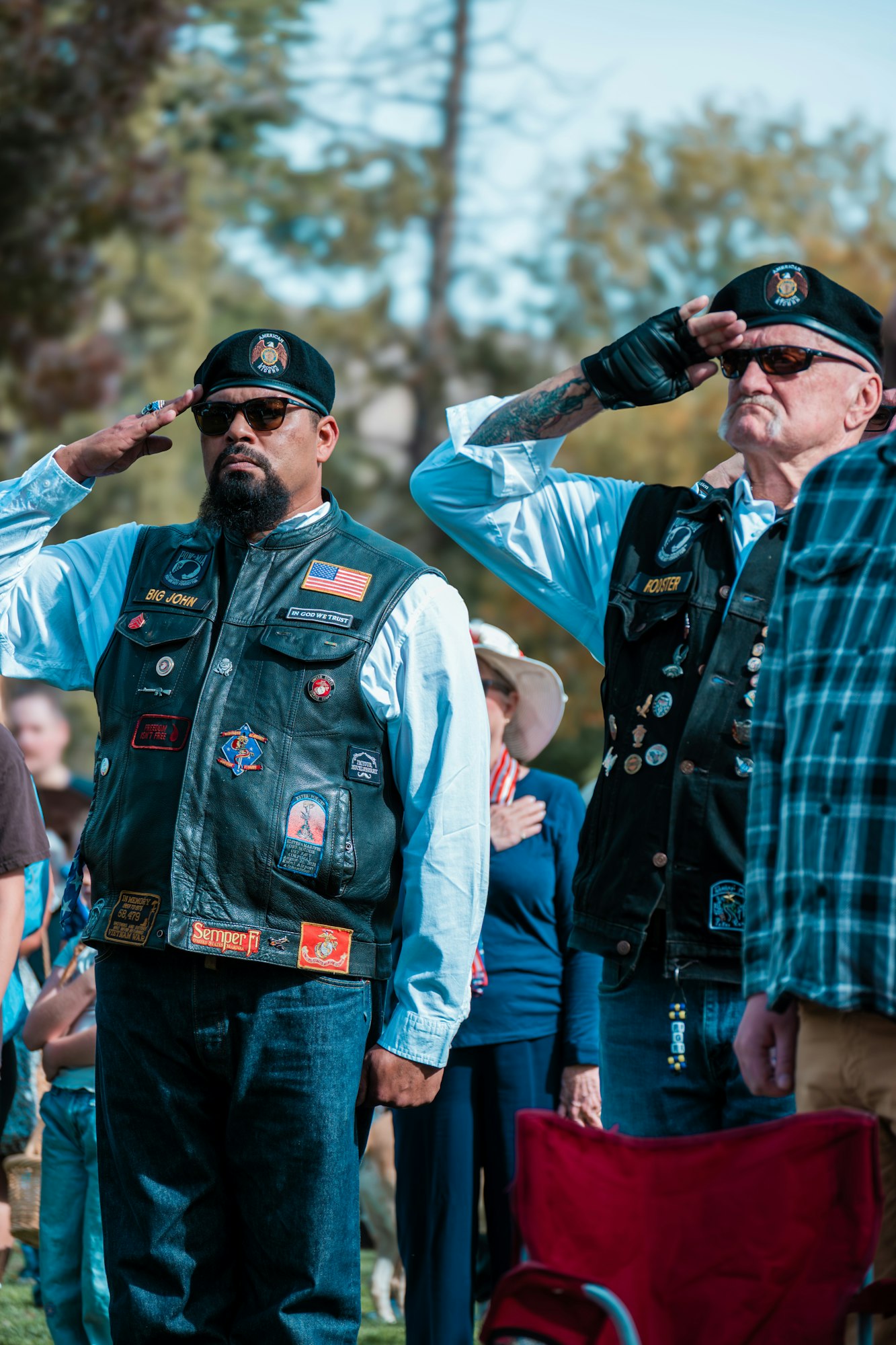 Two men in vests and hats salute outdoors, with patches and flags on their vests. Other people and trees in the background.