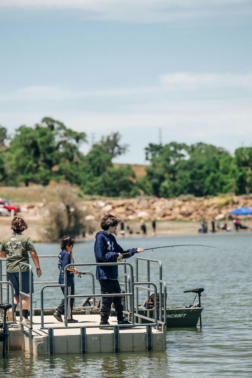 Three people fishing off a dock, trees in the background, sunny day.