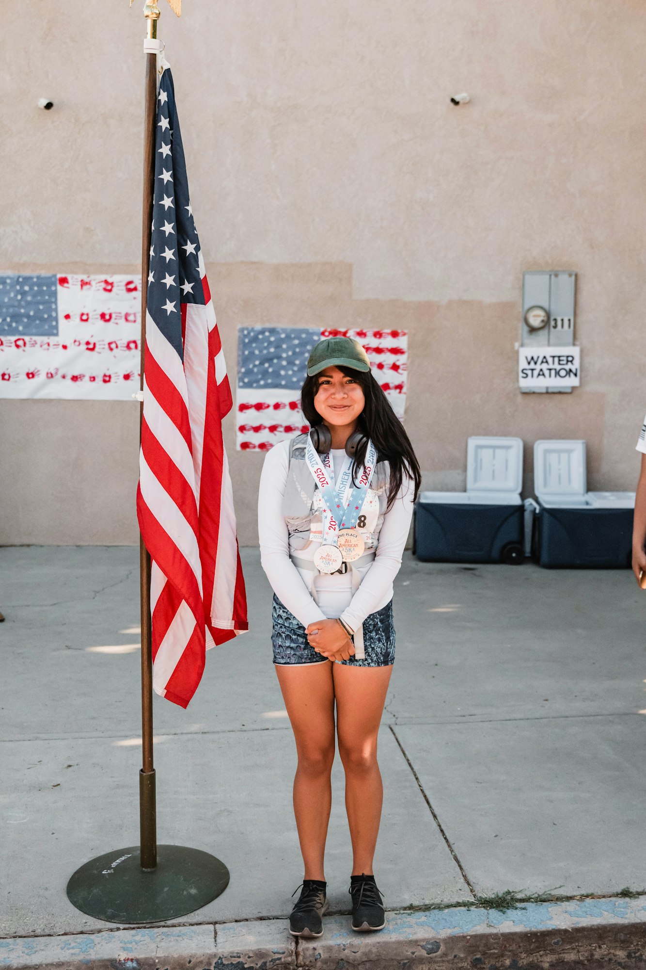 A person wearing medals stands beside a U.S. flag with coolers and a "Water Station" sign in the background.