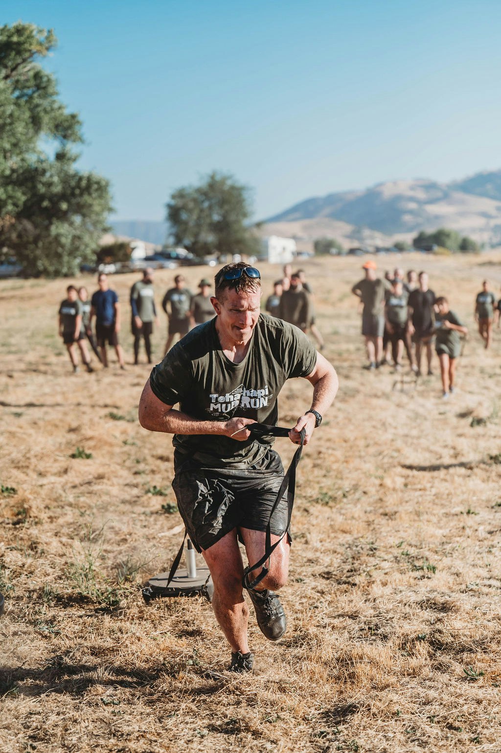 A person pulling a weighted sled during an outdoor event, with a group of people watching in the background.