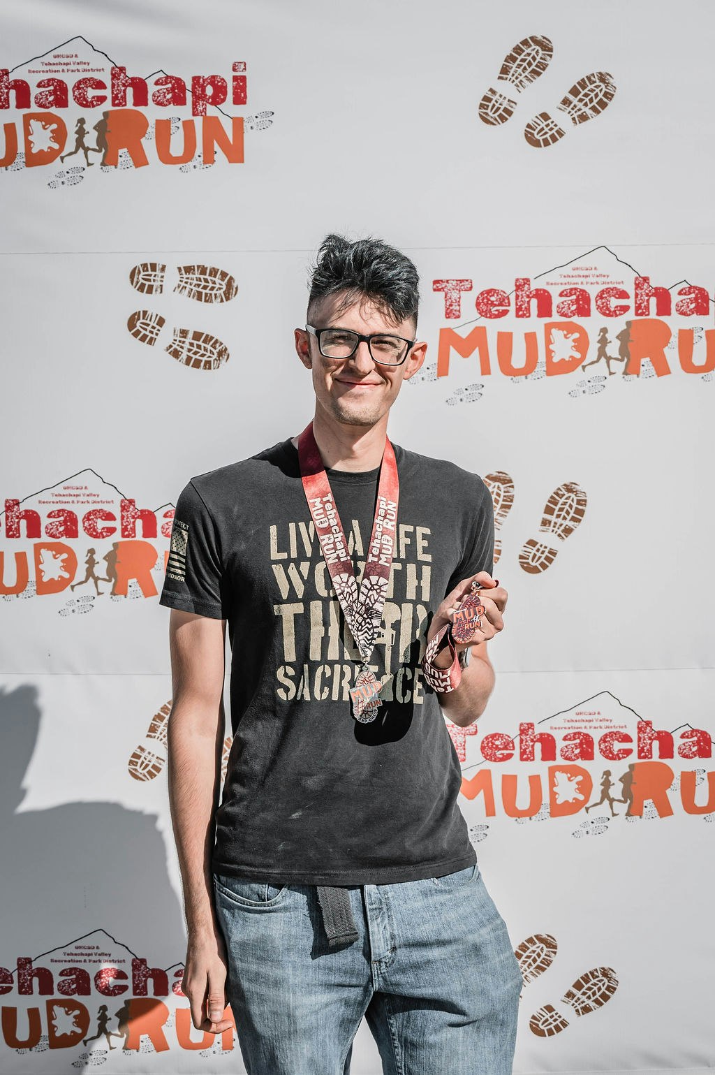 A person with glasses holds medals, standing in front of a "Tehachapi Mud Run" backdrop.