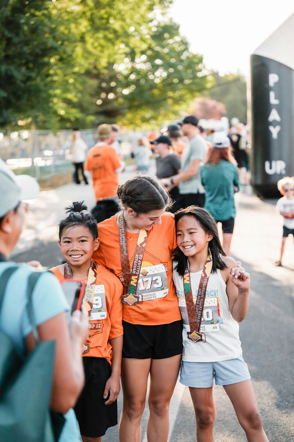 Three smiling kids wearing race bibs and medals, posing for a photo at an outdoor event.
