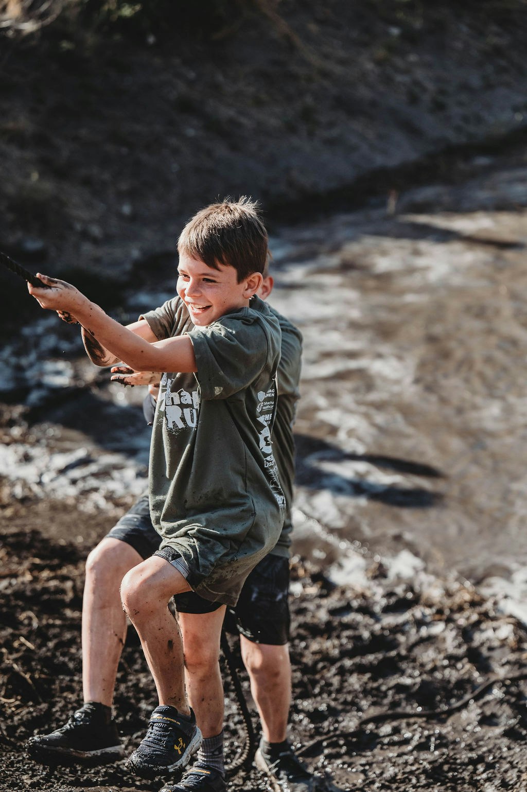 Two kids playing and pulling on a rope in a muddy outdoor setting, smiling and having fun.