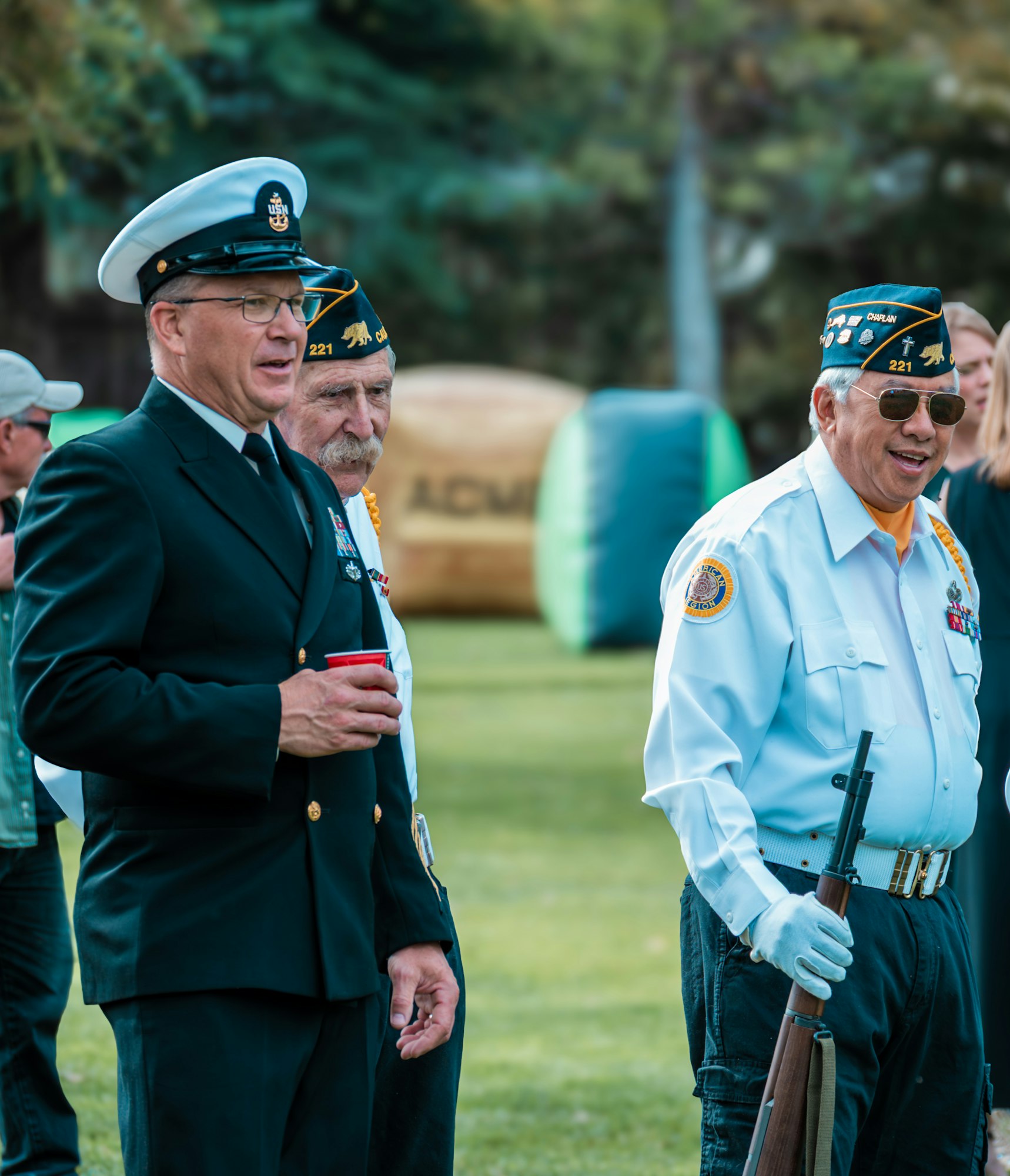 Three men in military uniforms at an outdoor event, one holding a rifle.