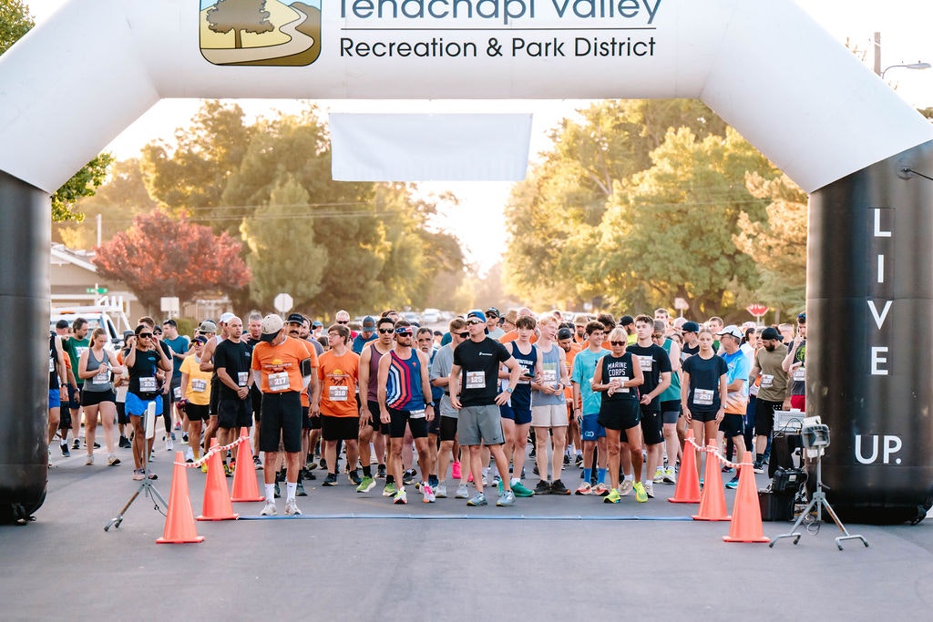 A group of runners is at the starting line of a race under an archway in a park setting.