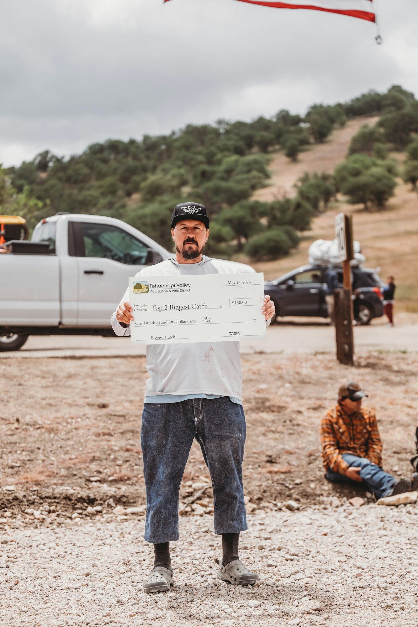 A person standing outdoors holding a large novelty check for $150, with a truck and a person sitting in the background.