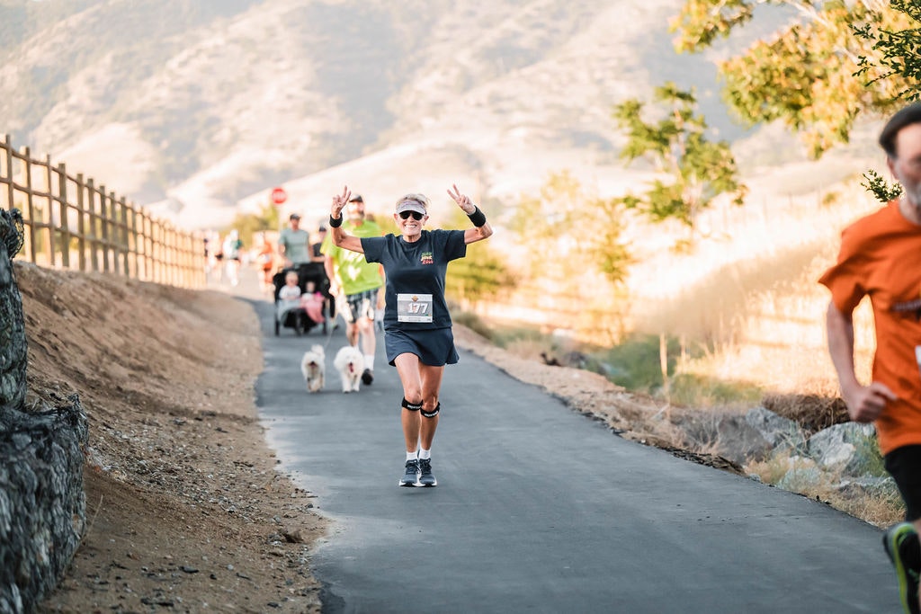 A group of people running on a scenic path, with a focus on a smiling runner in front wearing a race number bib.