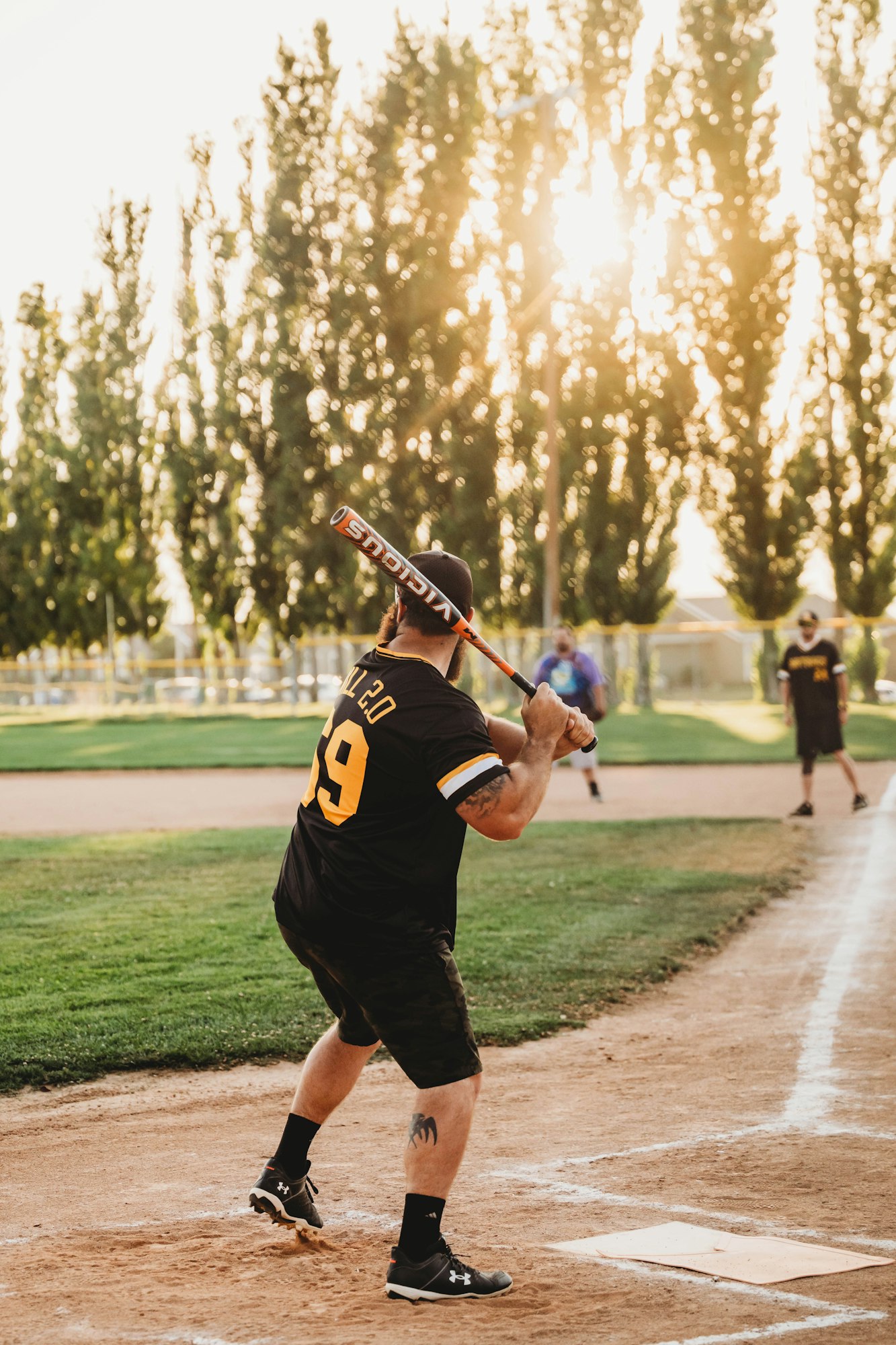 A player swinging a baseball bat on a field at sunset, with another player in the background.