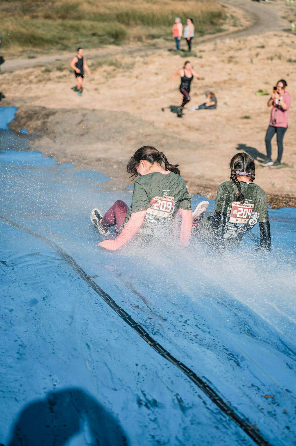 Two people slide down a wet, blue tarp in an outdoor race event, with others watching nearby.