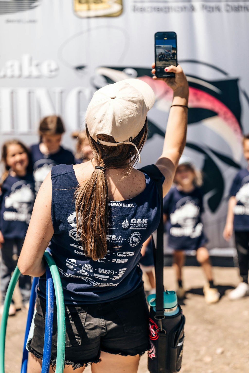Woman taking a selfie with kids in the background at an outdoor event.