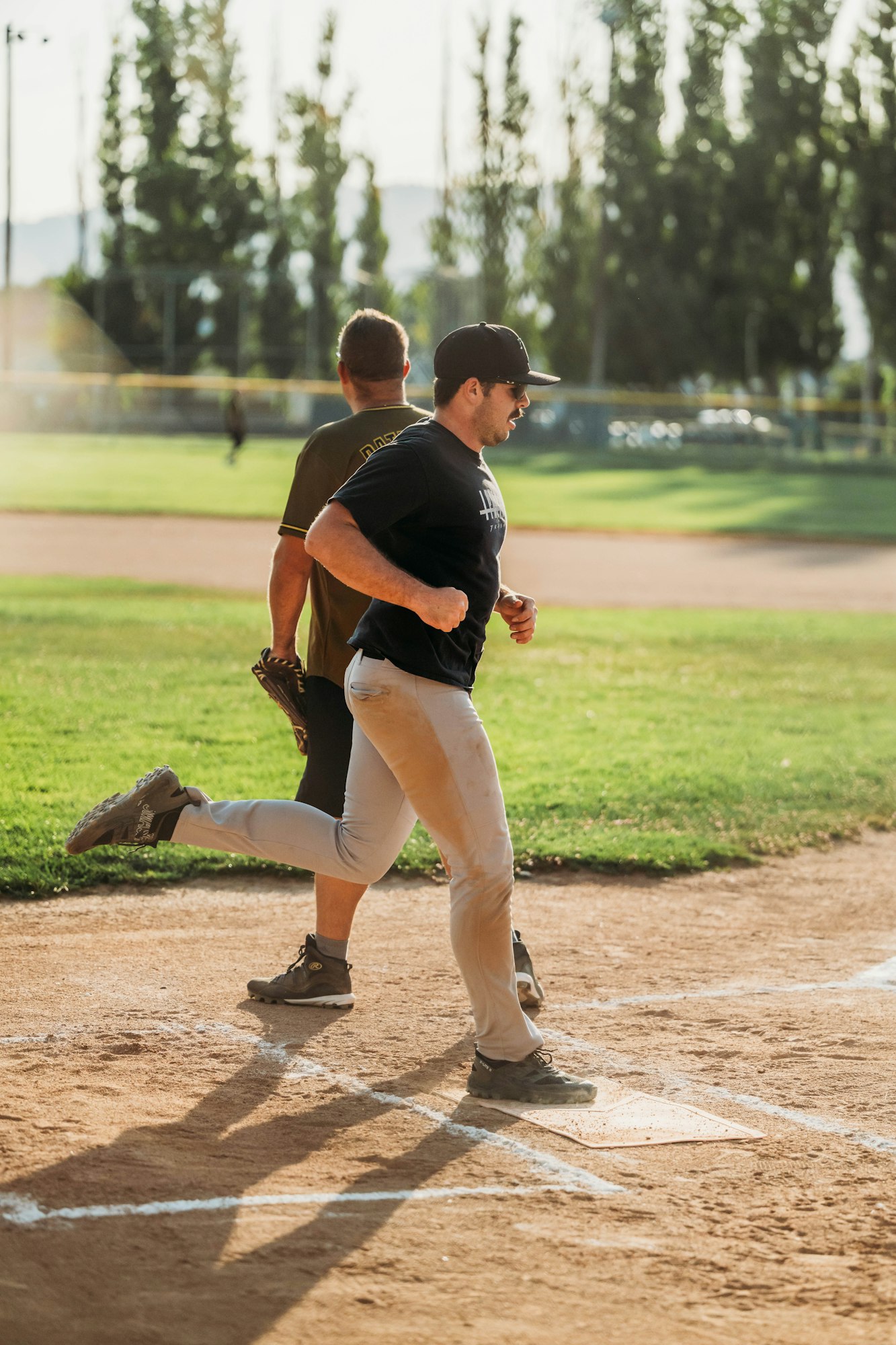 Two baseball players on the field, one running and the other watching, with trees in the background.