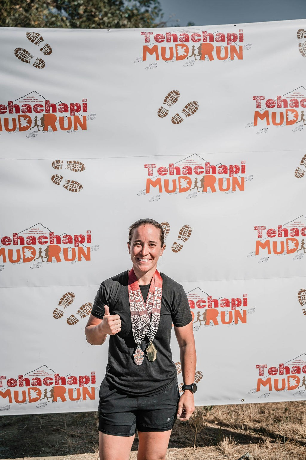 Person smiling, wearing medals, in front of "Tehachapi Mud Run" backdrop, giving a thumbs up.