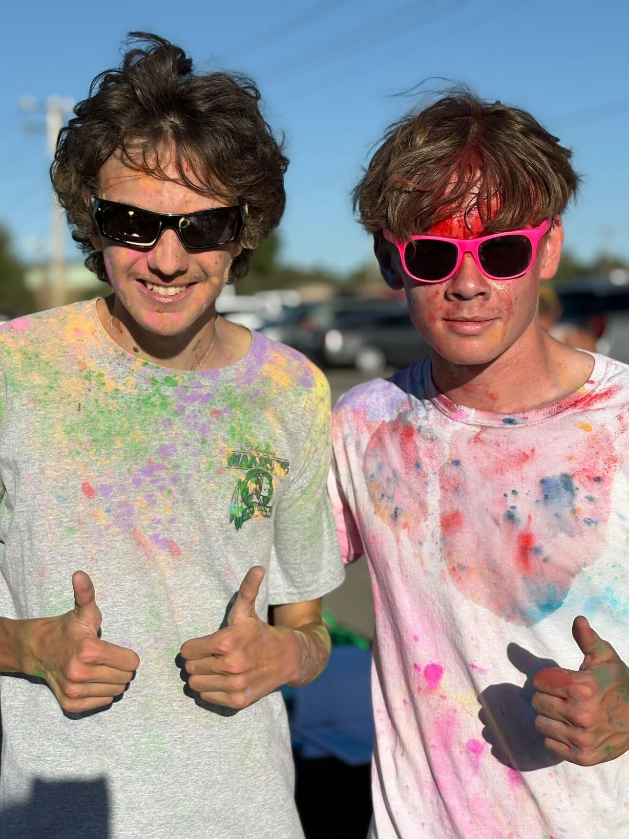 Two people wearing sunglasses and colorful, paint-splattered shirts give thumbs up.