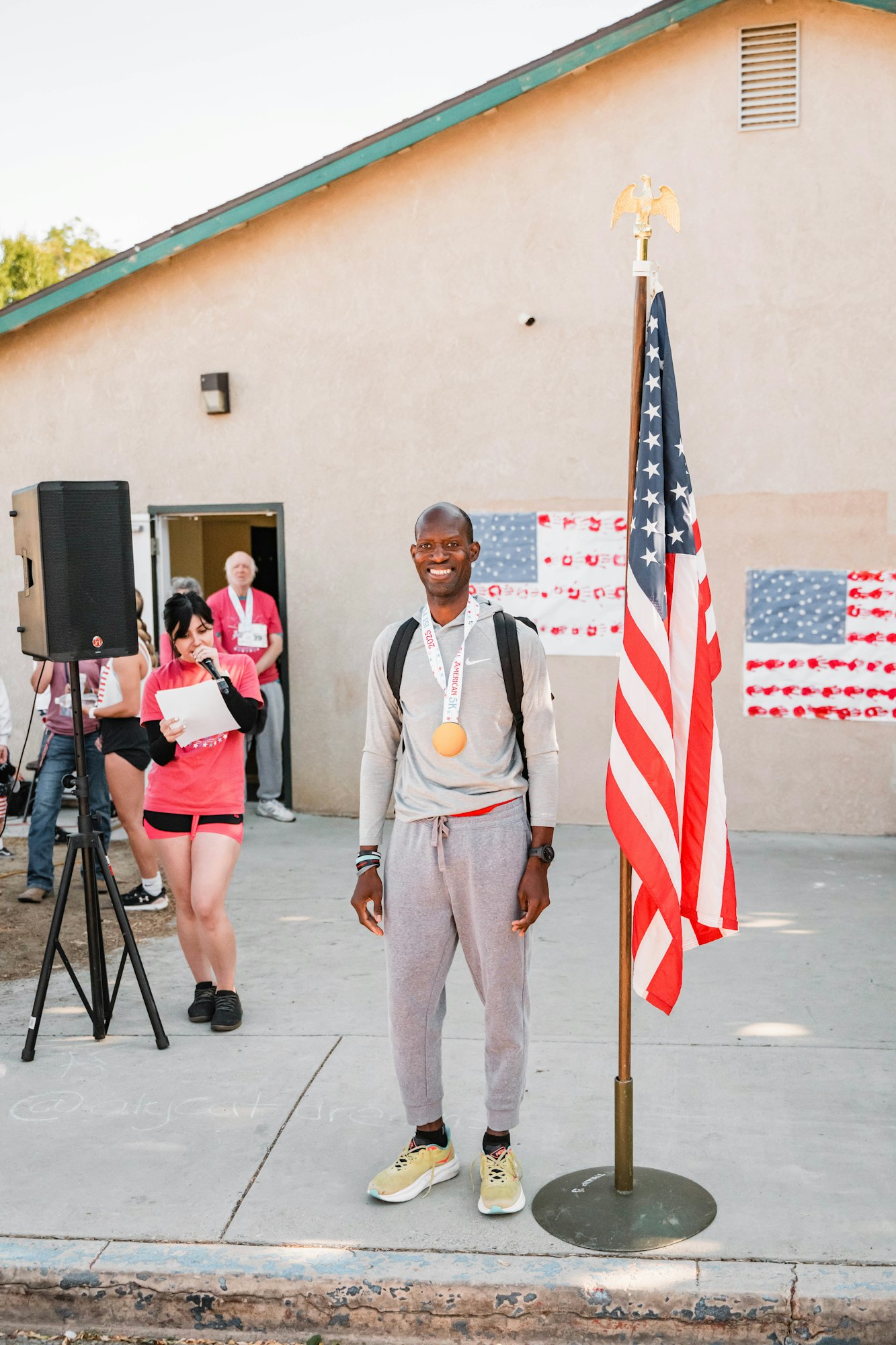 A person stands smiling next to an American flag wearing a medal, with people in the background at an outdoor event.