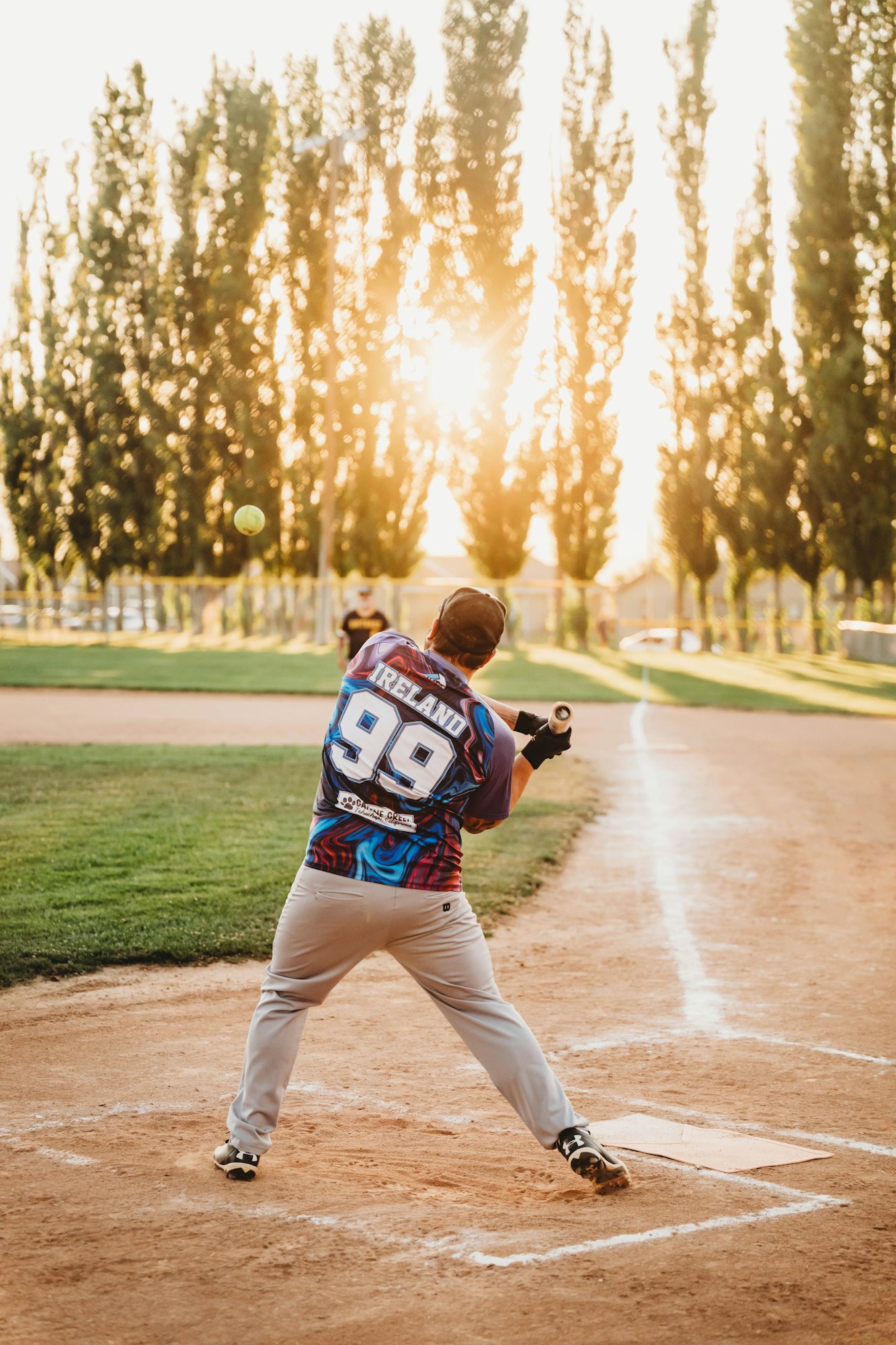 A baseball player swinging a bat with the ball in mid-air, bathed in sunlight with trees in the background.