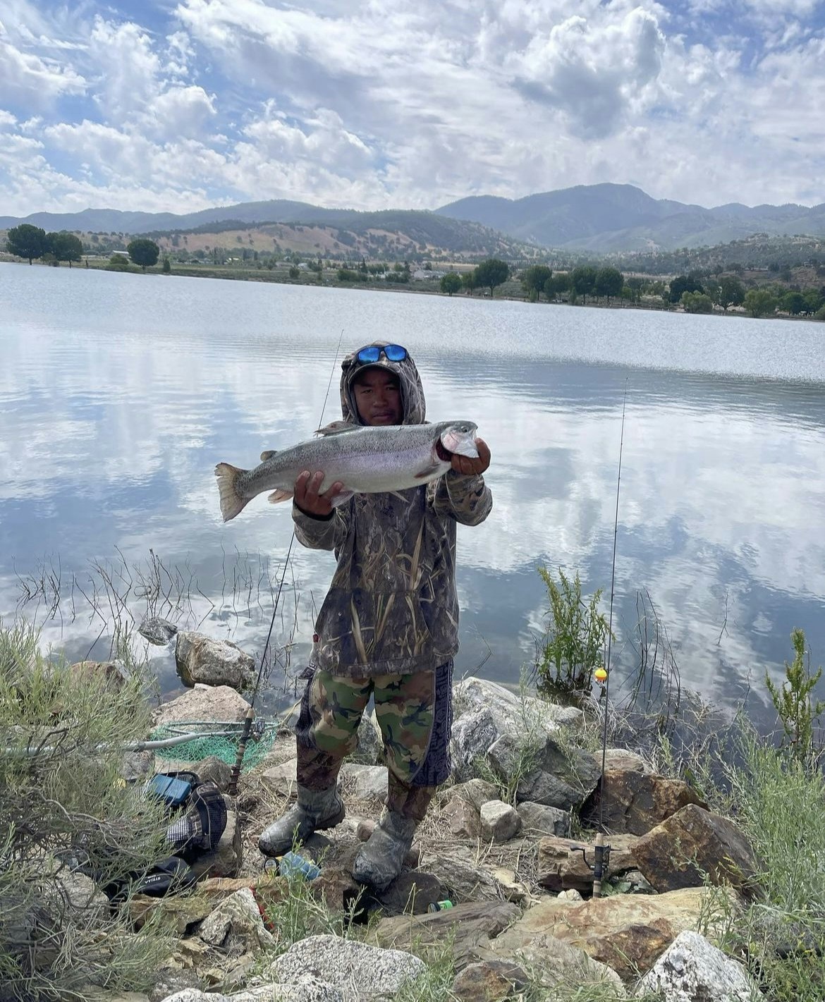 A fisherman holding a trout by a lake, with fishing gear, camo clothing, and mountains in the background.