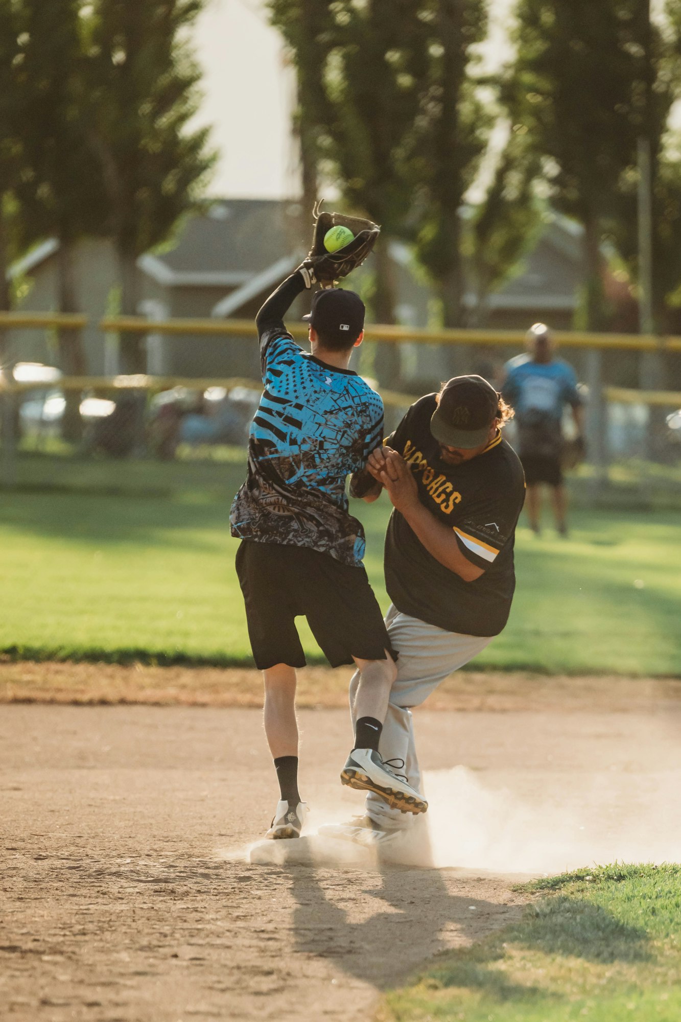 Two people playing softball, one catching a ball above his head and the other sliding into base, creating a cloud of dust.