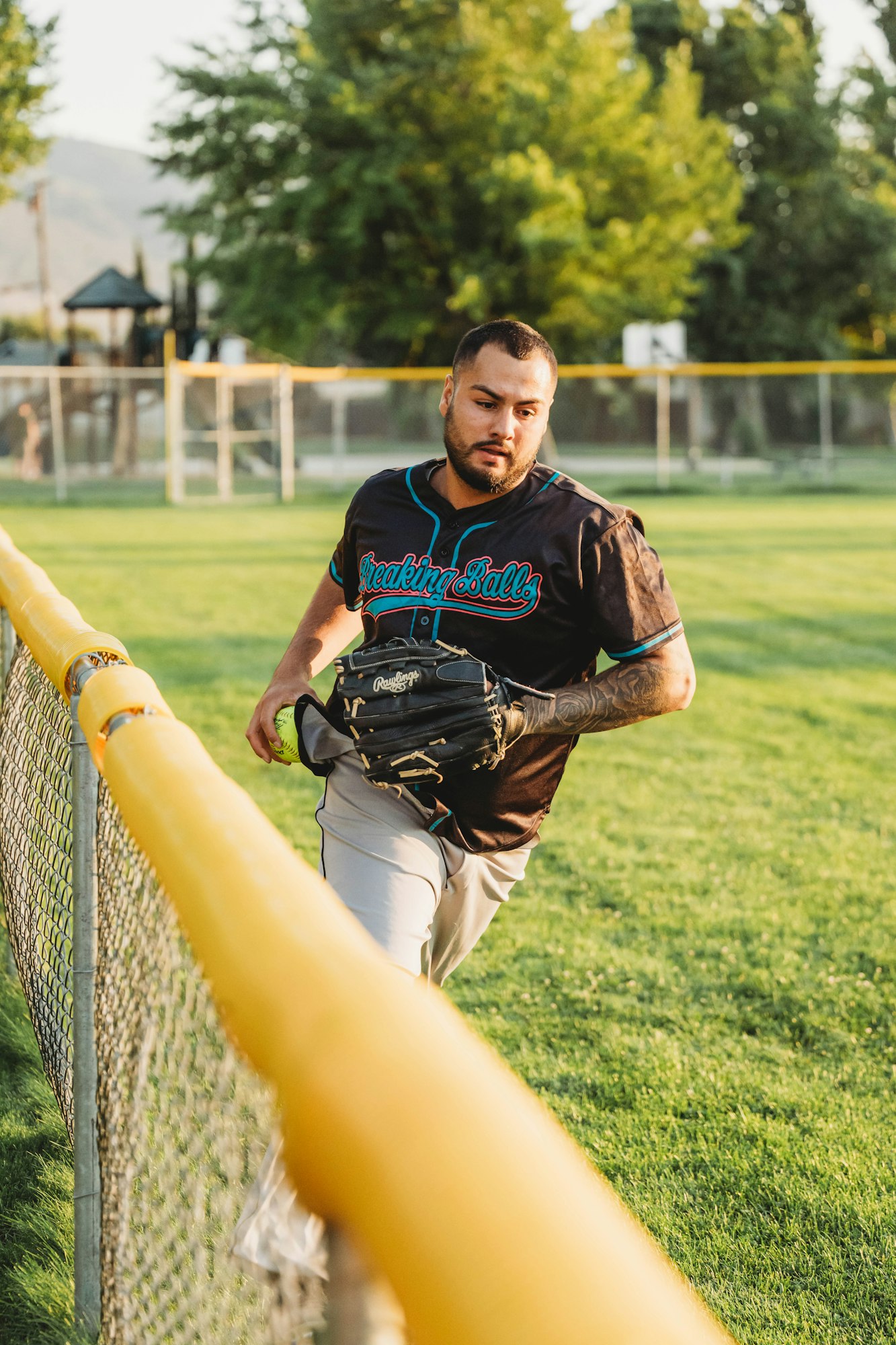 A baseball player holding a glove, near a fence on a sunny day.