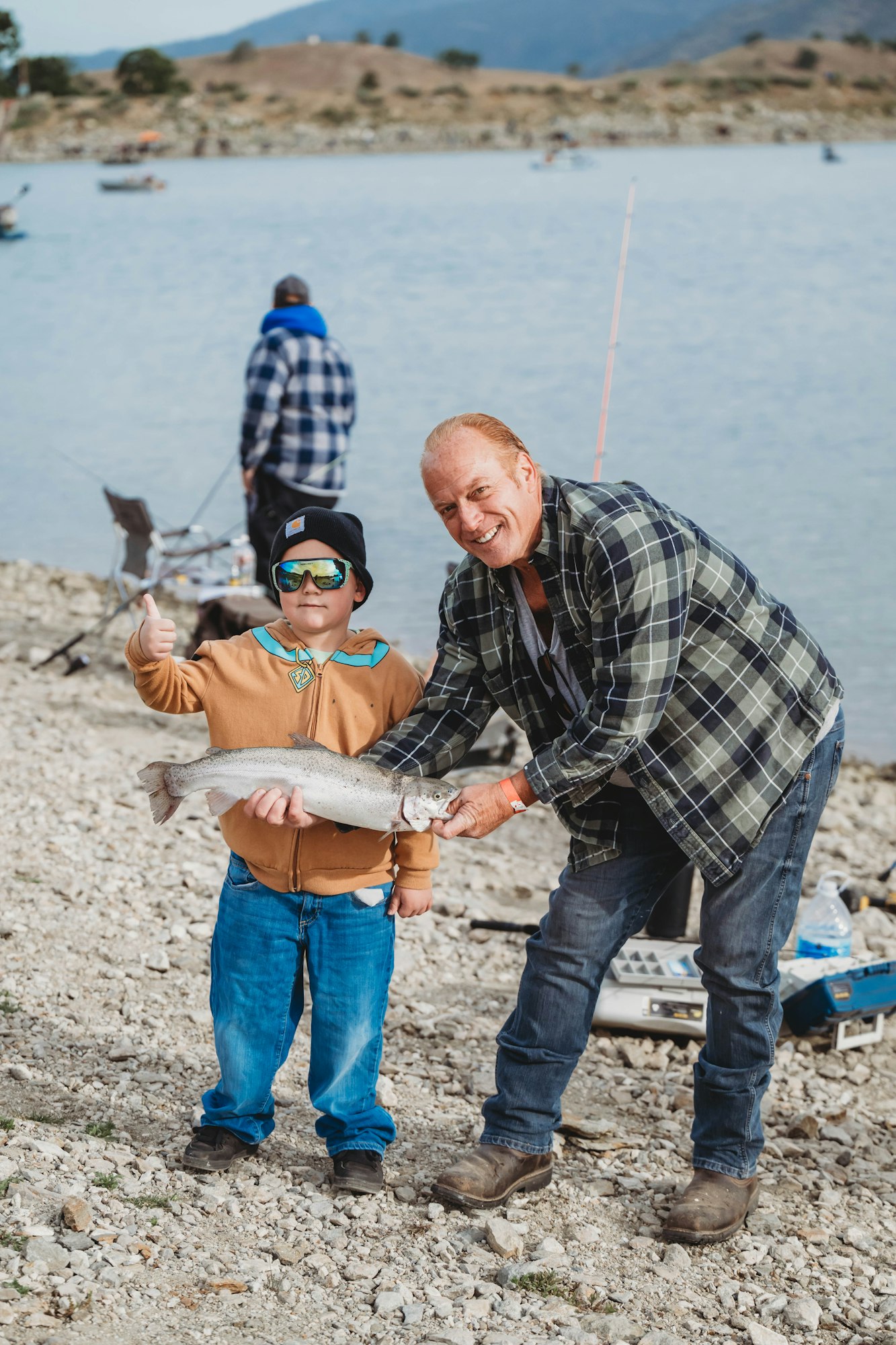 A child and adult smiling, holding a large fish by a lakeside, with fishing gear in the background.