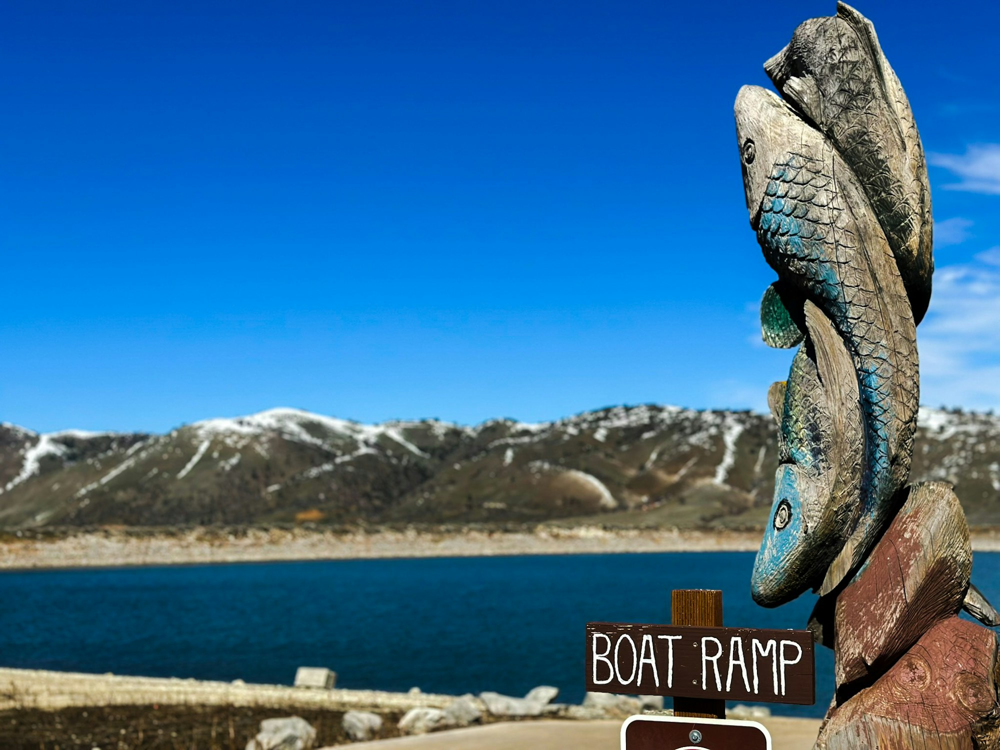 Carved fish sculpture by a lake with mountains in the background, next to a "BOAT RAMP" sign.