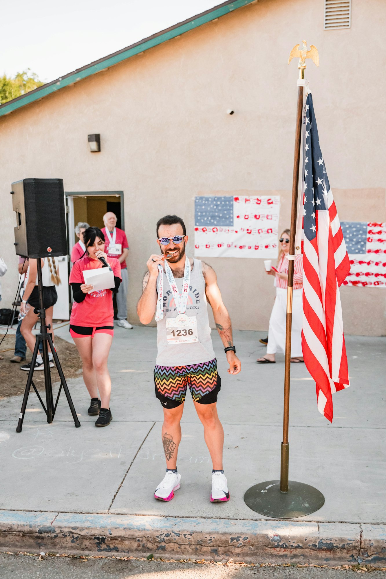 A man poses with a medal near a U.S. flag, wearing a race bib and colorful shorts, surrounded by people and an outdoor speaker.