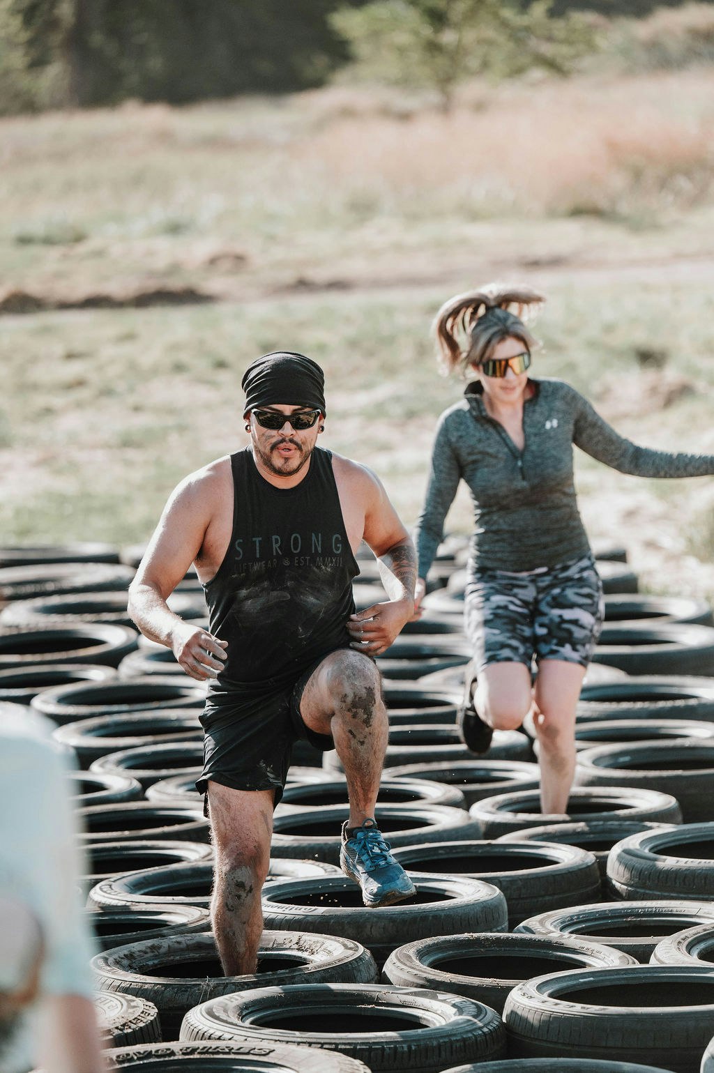Two people running on a tire obstacle course in sporty attire, outdoors.