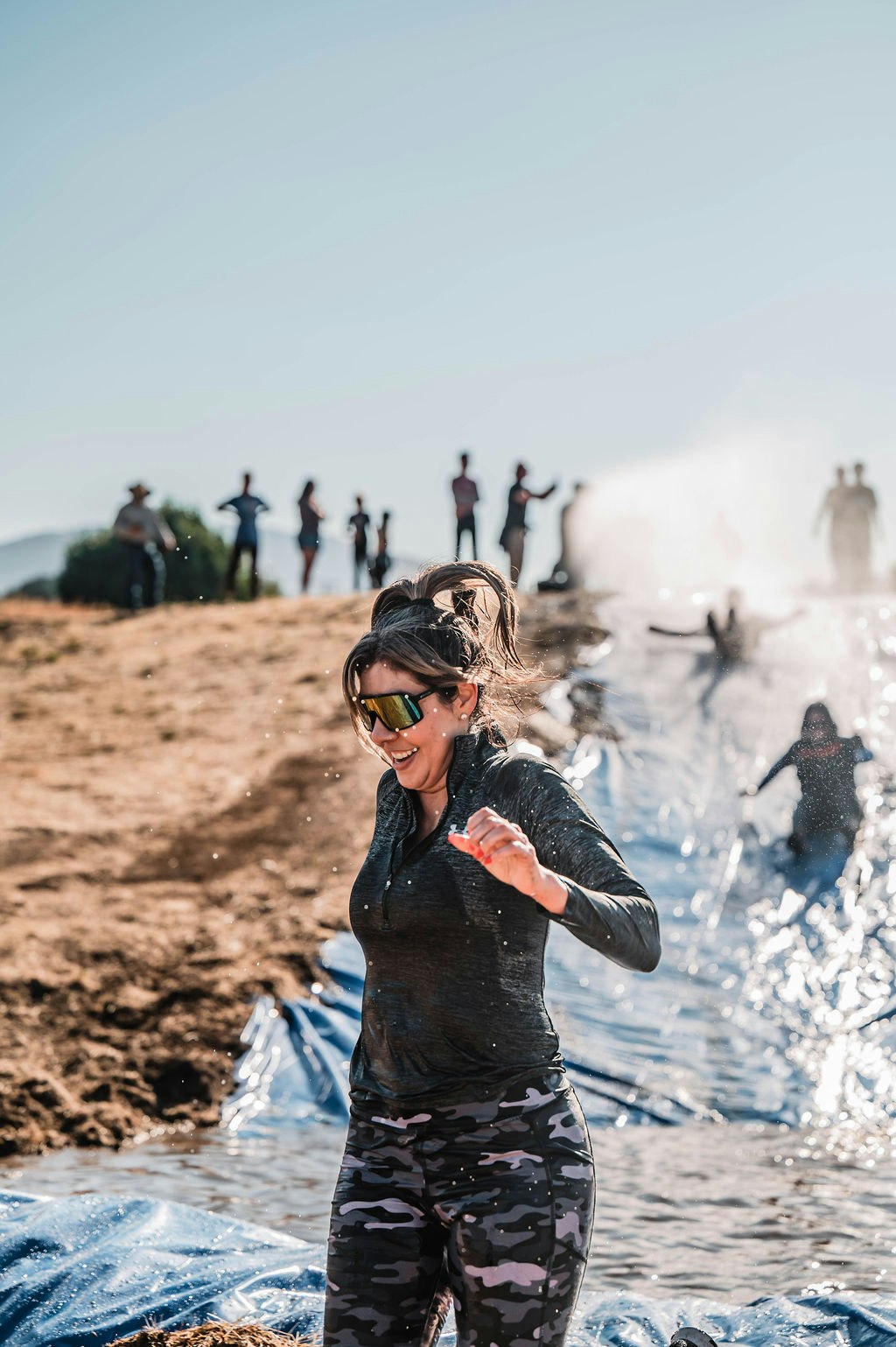 A woman in sunglasses enjoys sliding down a water slide outdoors, with others in the background.