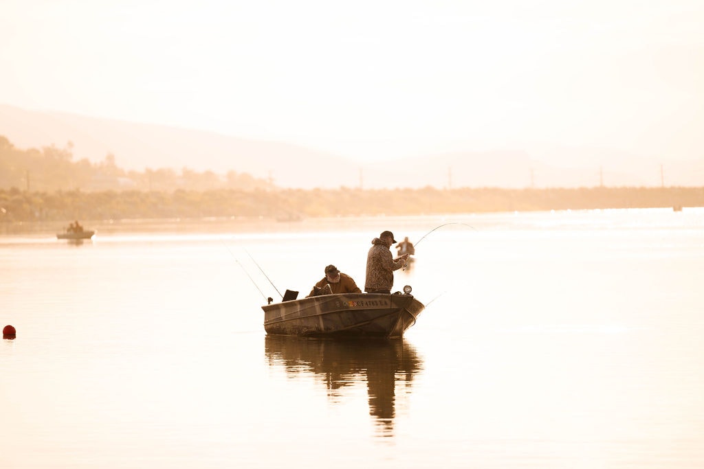 Two people fishing from a boat on calm water with a hazy, golden sunset.