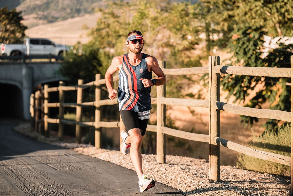 A man running on a sunny path beside a wooden fence, wearing a striped tank top, sunglasses, and a headband.