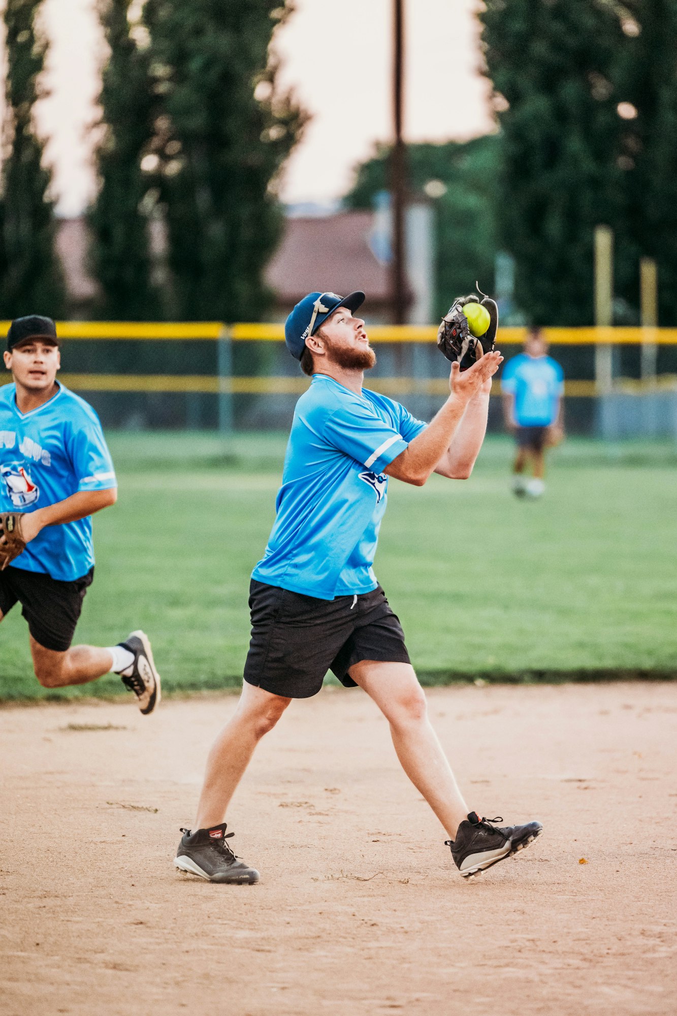 Two softball players in blue uniforms, one catching a ball, the other in the background, on a dirt field.