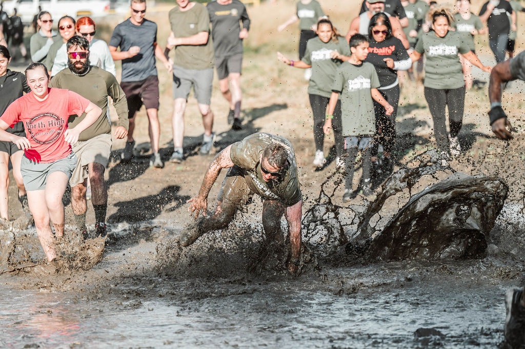 People running through muddy water, some splashing, during an outdoor obstacle event.