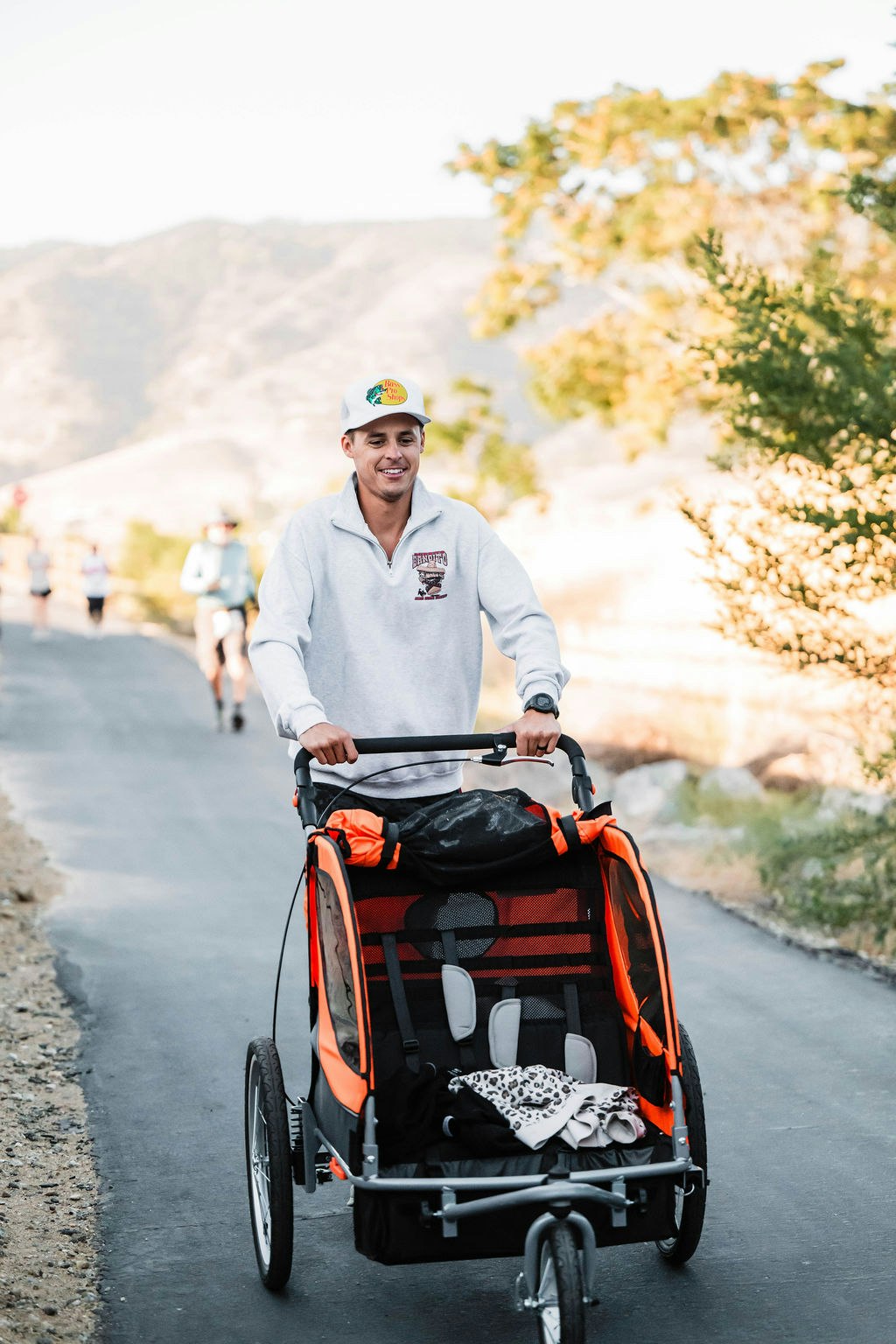 A person pushing a bright orange stroller on a path, outdoors, wearing a cap and sweatshirt.
