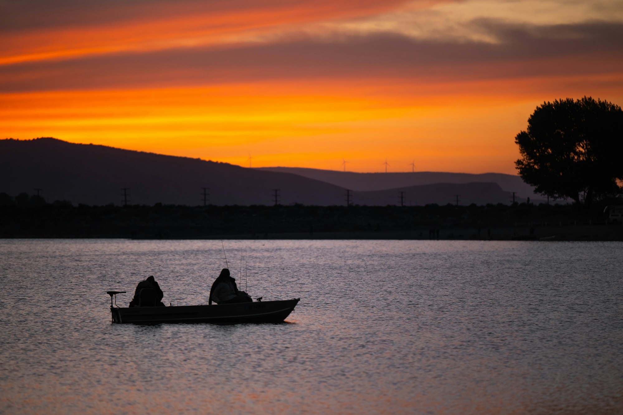 Silhouetted fishermen in a boat at sunset, with hills and wind turbines in the background.