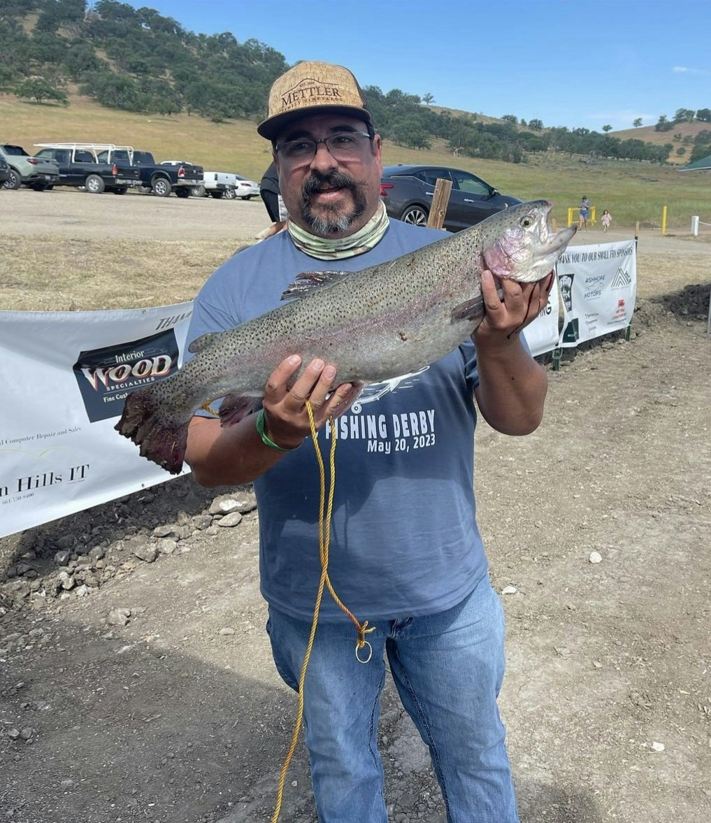 Man holding a large fish at a fishing derby event on May 20, 2023. Cars and hills in the background.