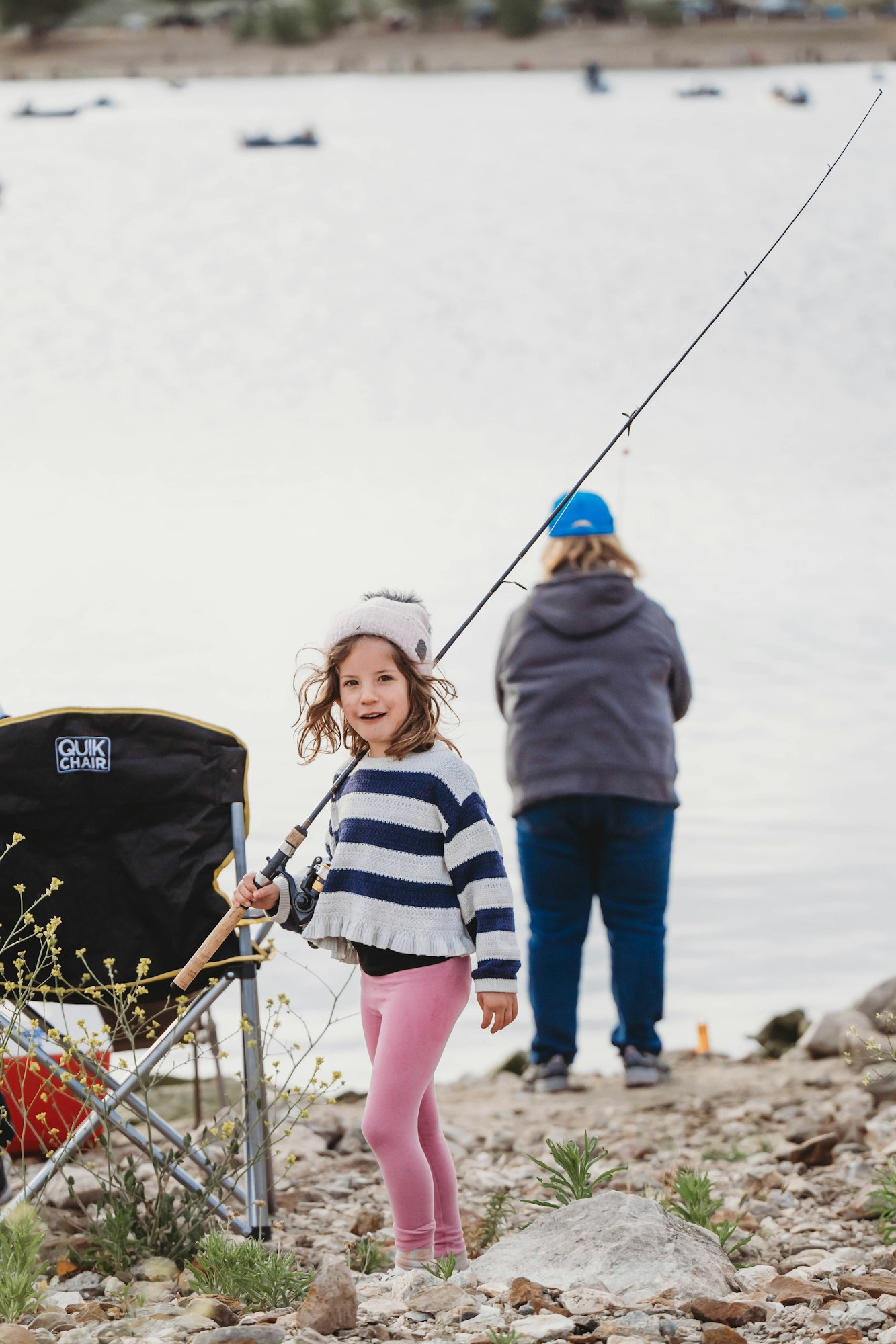 Child holding a fishing rod on a rocky shore, with an adult and water in the background.