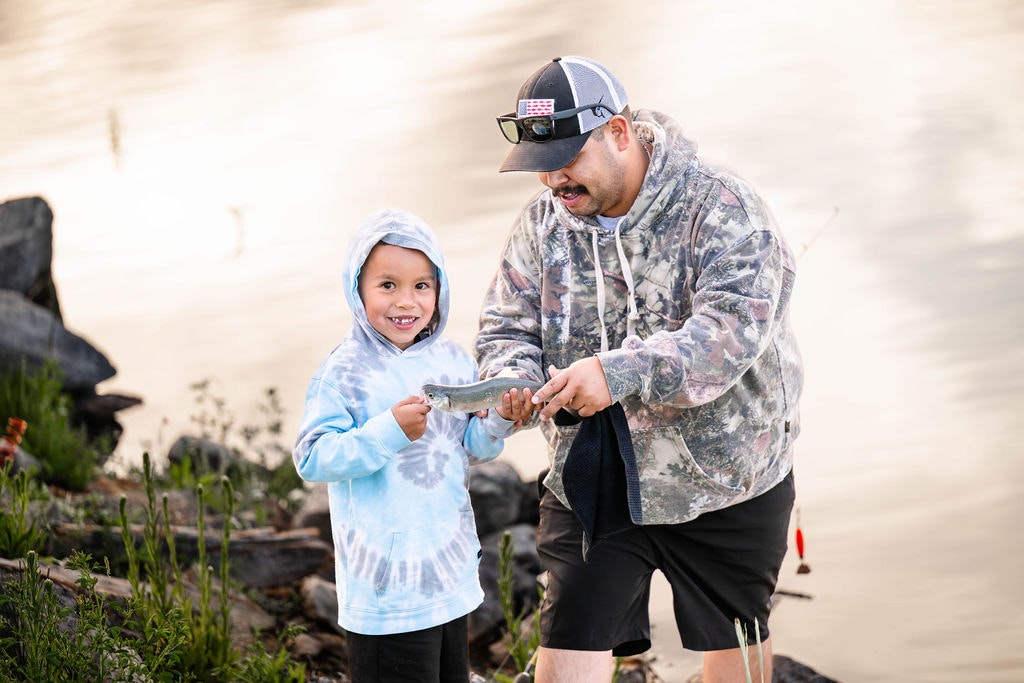 Adult and child holding a fish by a lake, smiling, with fishing gear in the background.