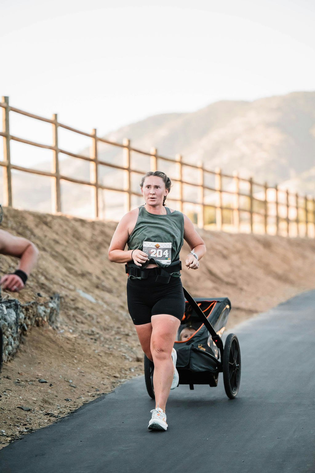A person running on a path, pushing a stroller with a child, wearing a race number.