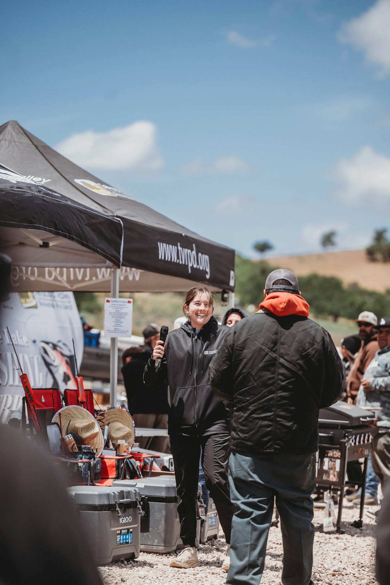 A person with a microphone smiles under a canopy, talking to a crowd outdoors. Camping gear is displayed around the area.