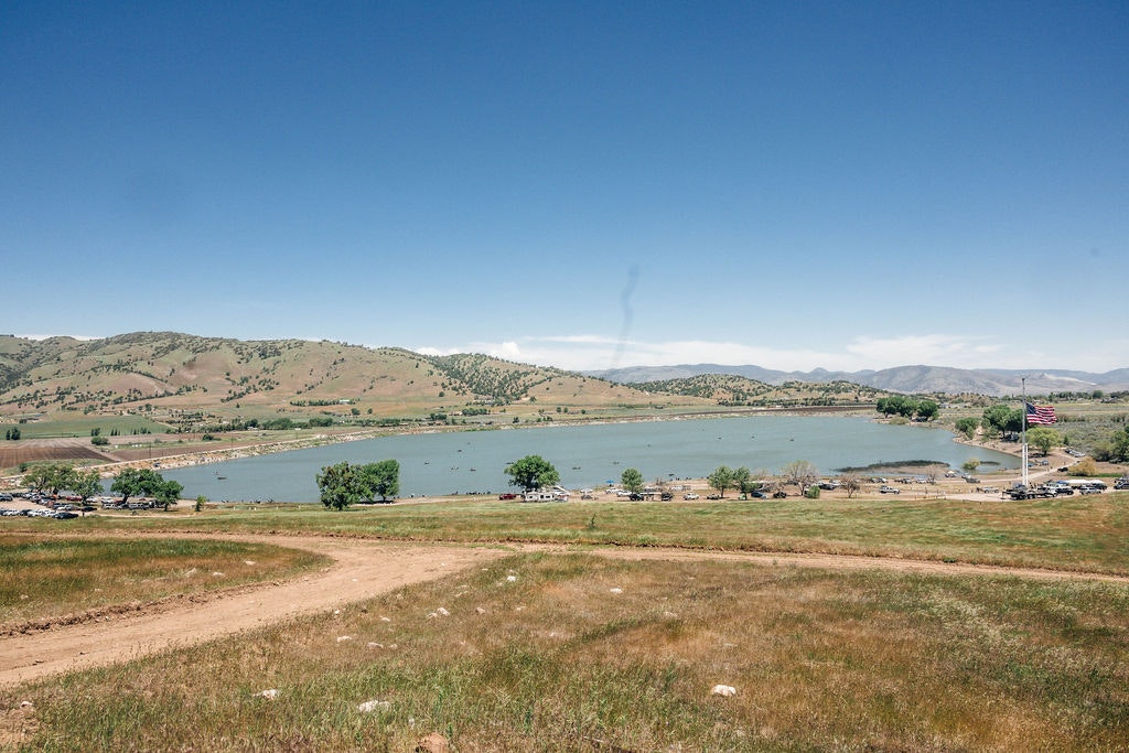 A lake surrounded by grassy hills, parked cars, a large American flag, and a clear sky.