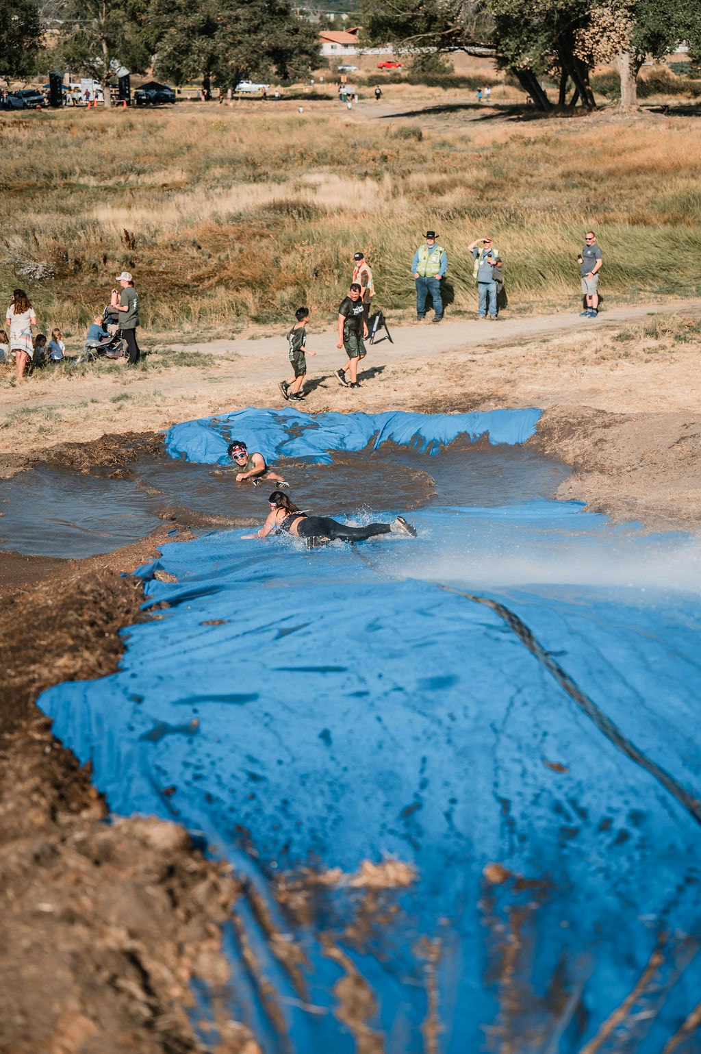 People sliding down a muddy hill covered with a blue tarp, with spectators watching in a grassy area.