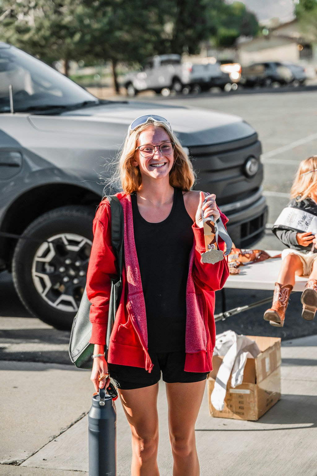 A smiling person wearing a red jacket holding keys and a water bottle, standing near a car and a table with a box.
