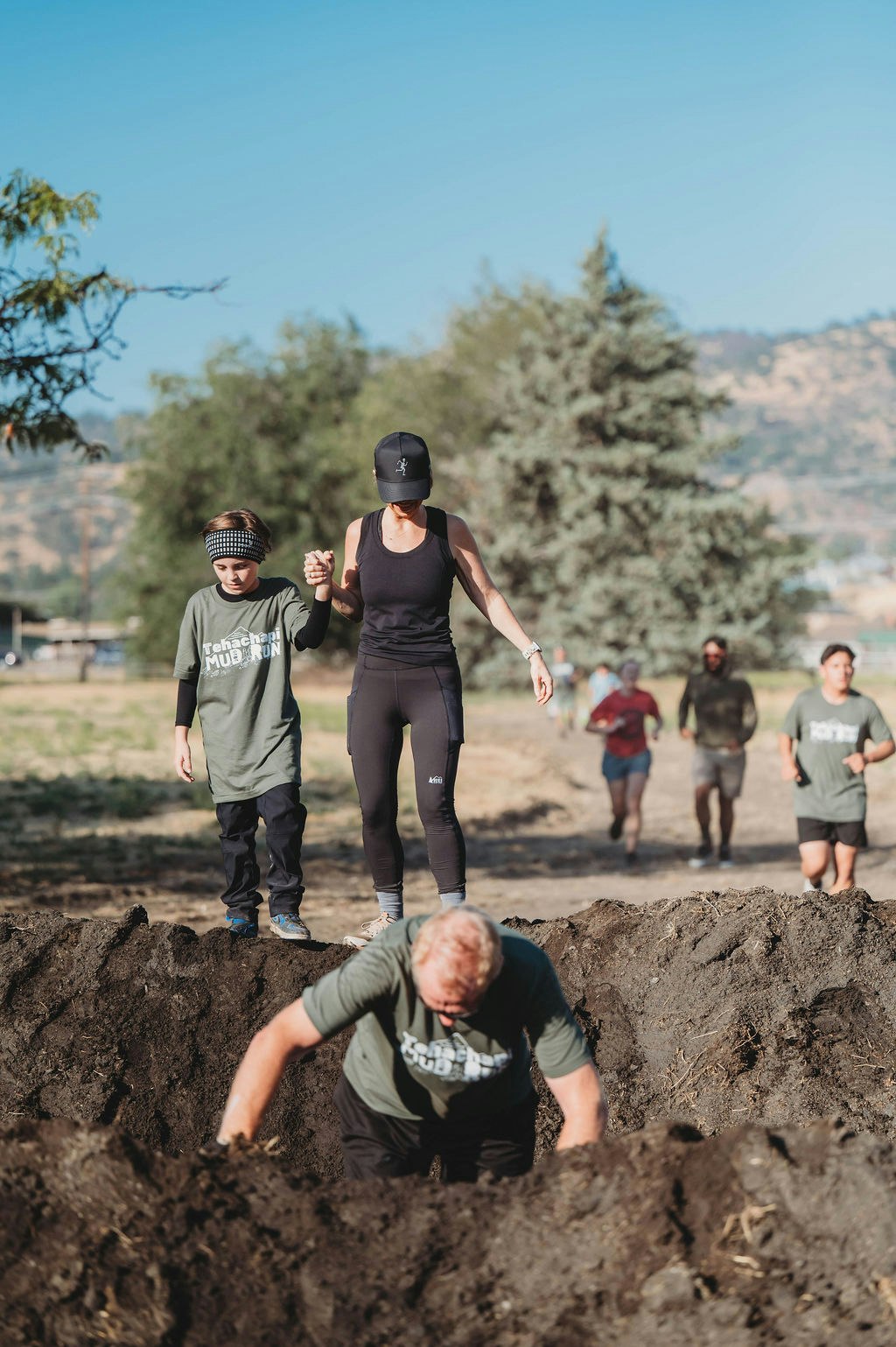 A group of people participating in a mud run event, with a woman and child holding hands, navigating over a dirt mound.