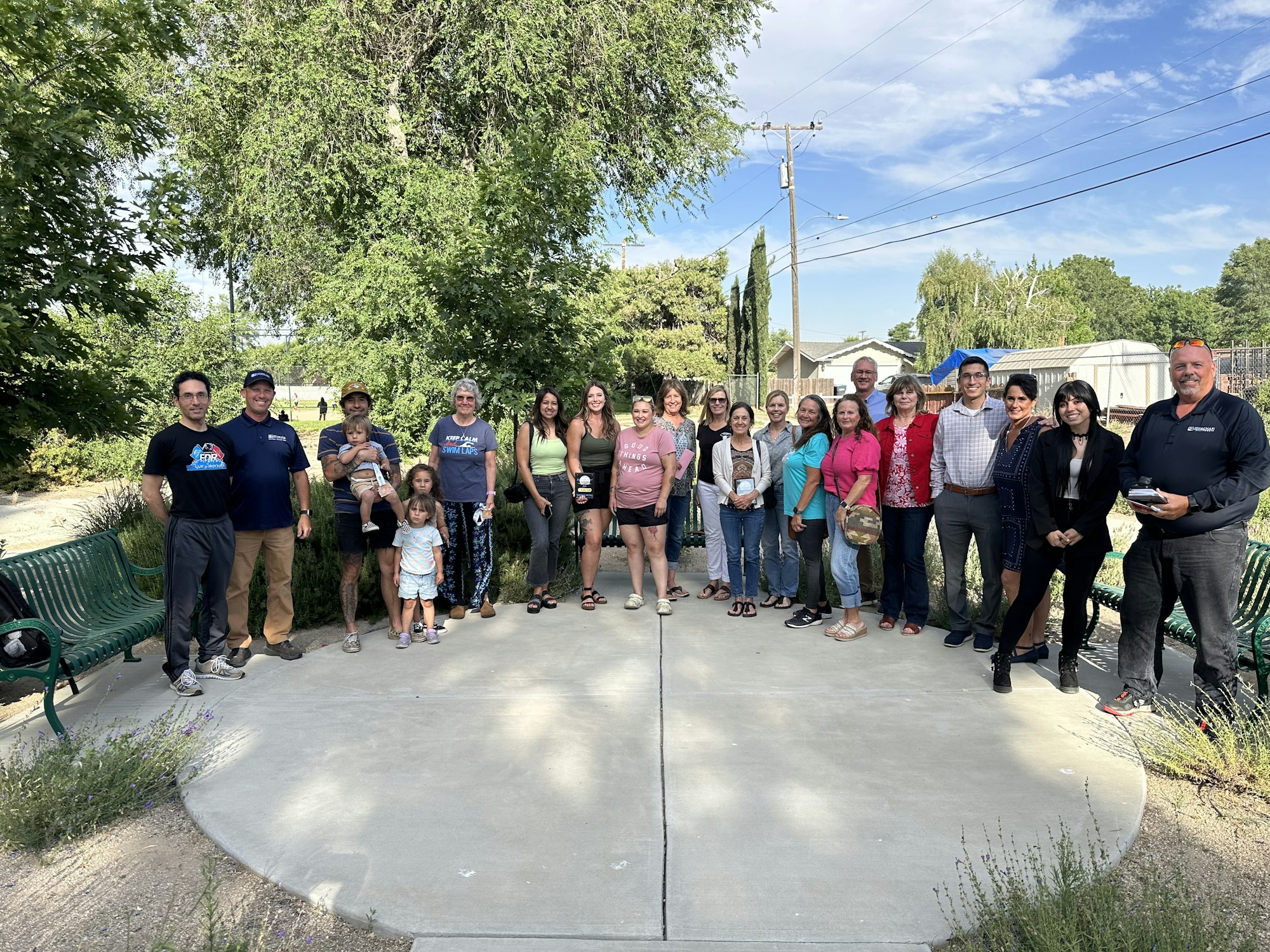 Group of people posing for a photo in an outdoor park setting with trees and a bench.