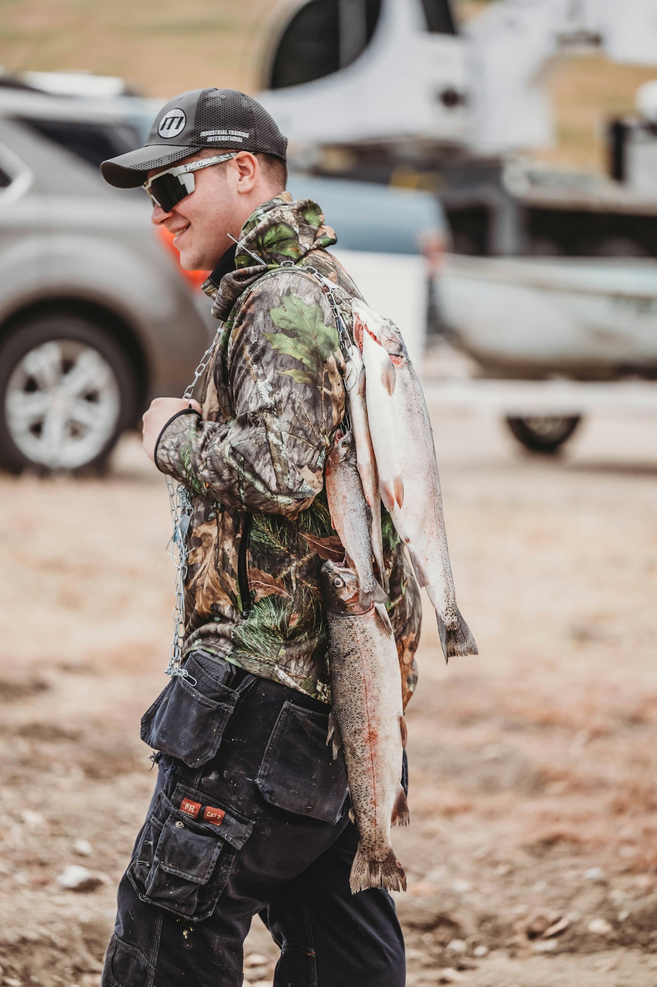 A person in camo clothing carries fish on a chain, with cars and equipment in the background.