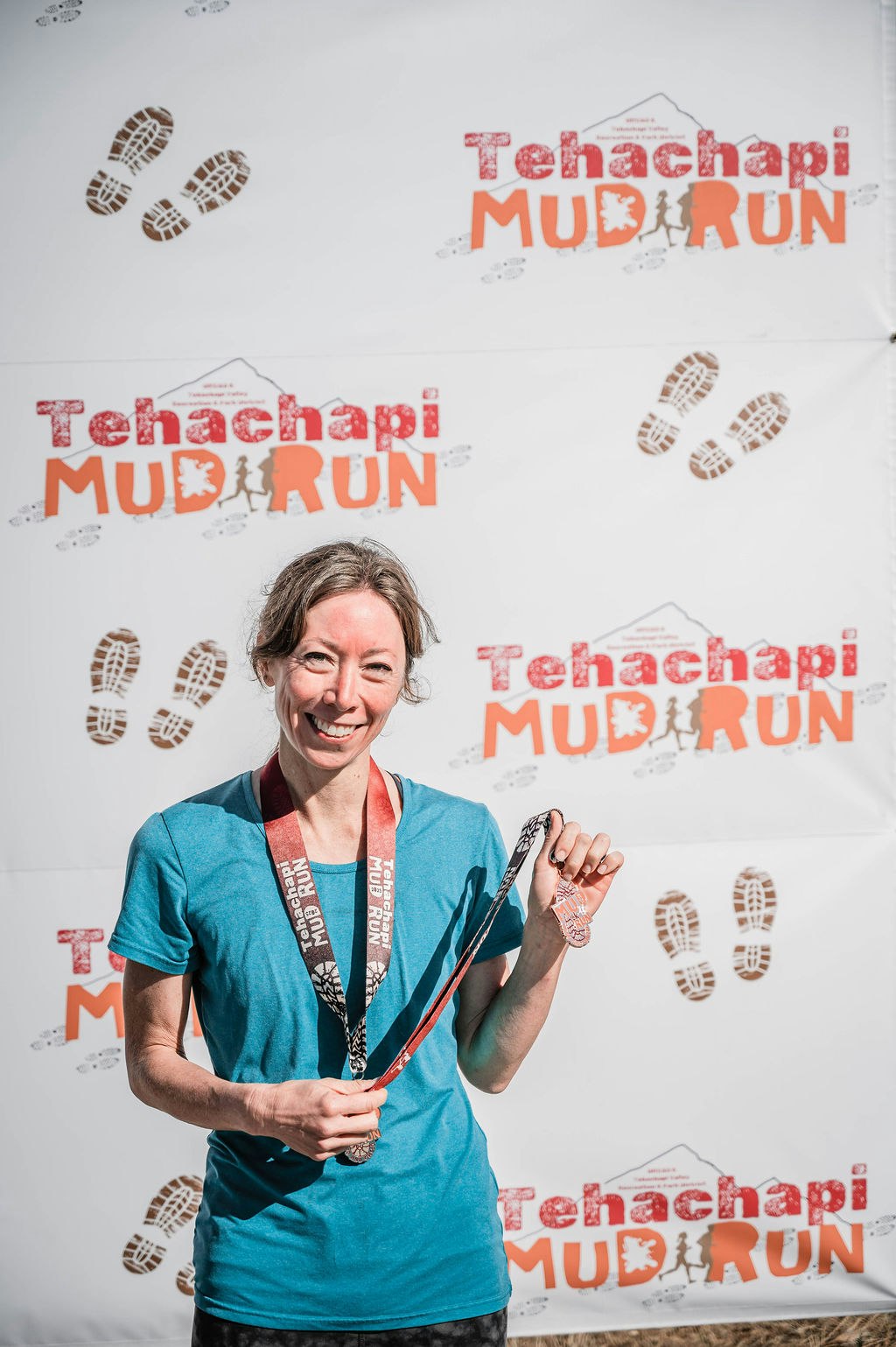 A person holding a medal stands in front of a "Tehachapi Mud Run" backdrop.