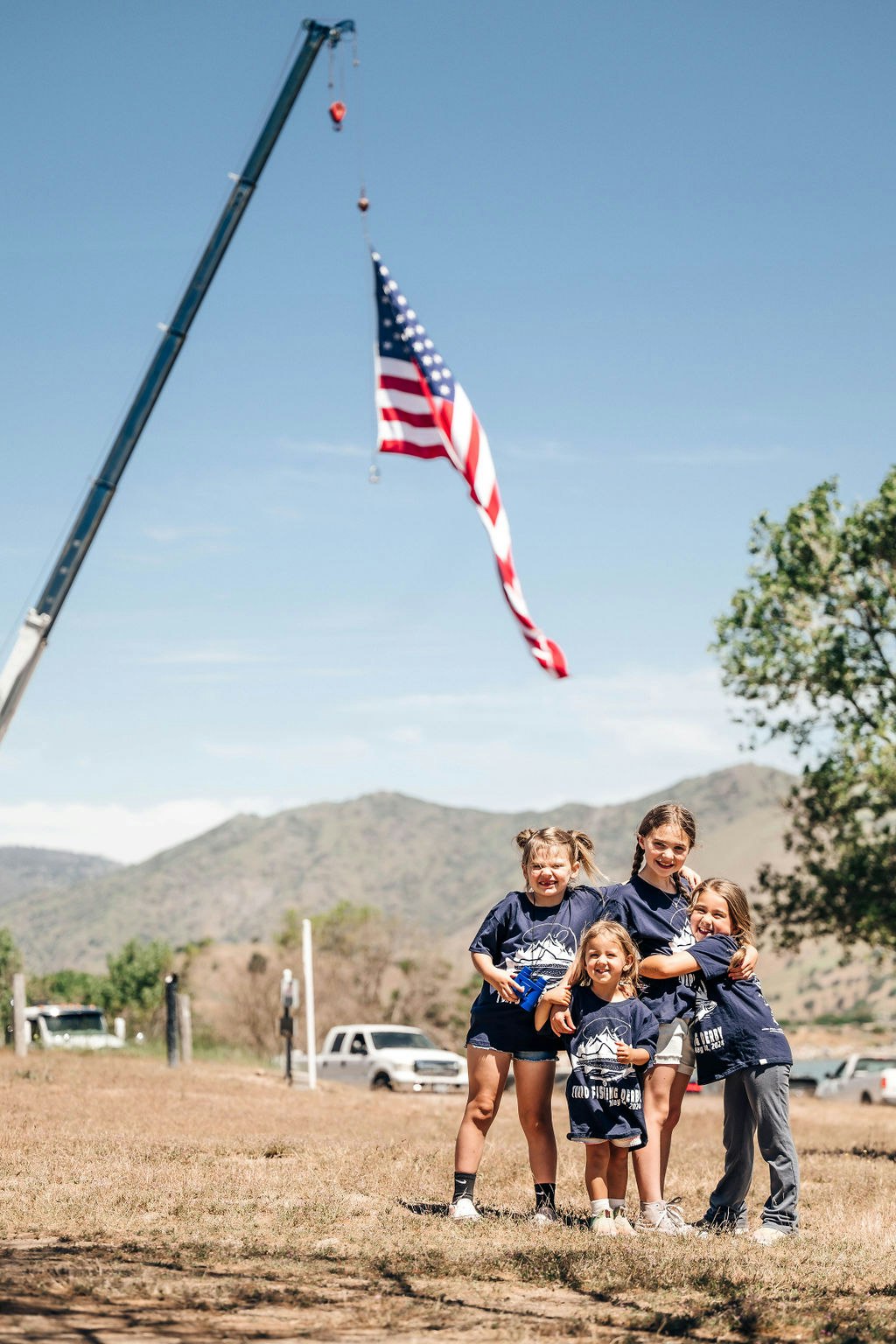 Group of smiling kids posing outdoors with a large American flag hanging from a crane in the background.