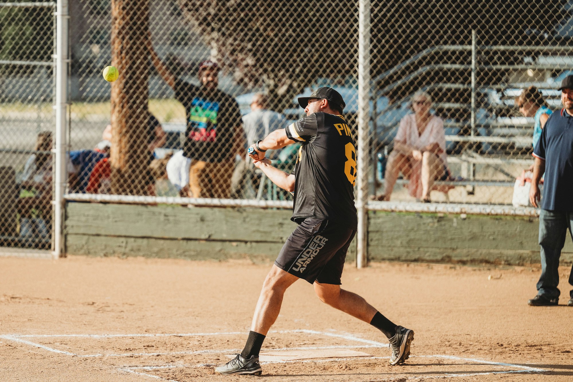 A softball player swinging at a pitch on a dirt field with spectators behind a fence.