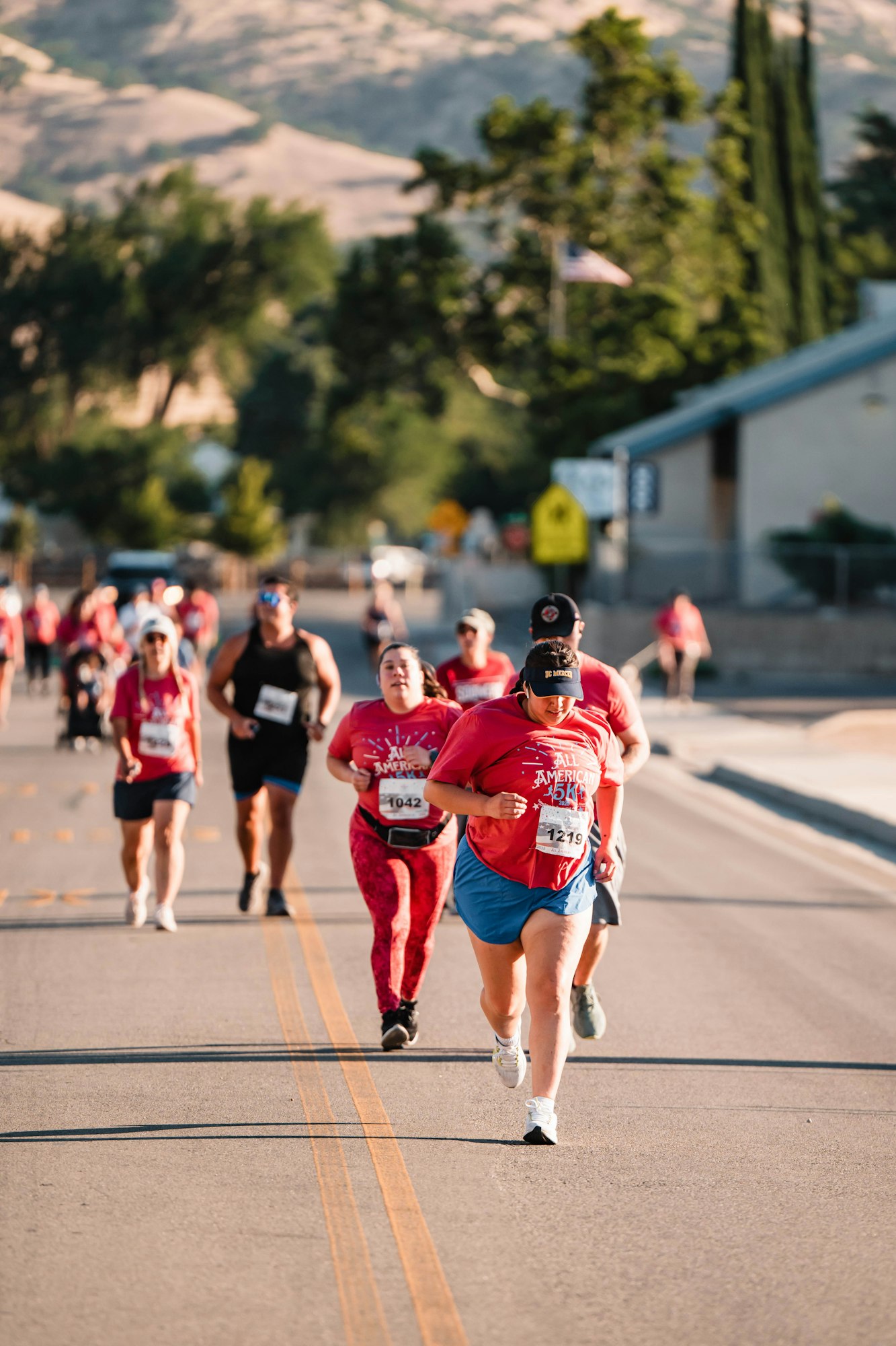 Runners participating in a road race, wearing bibs and athletic attire, with trees and hills in the background.